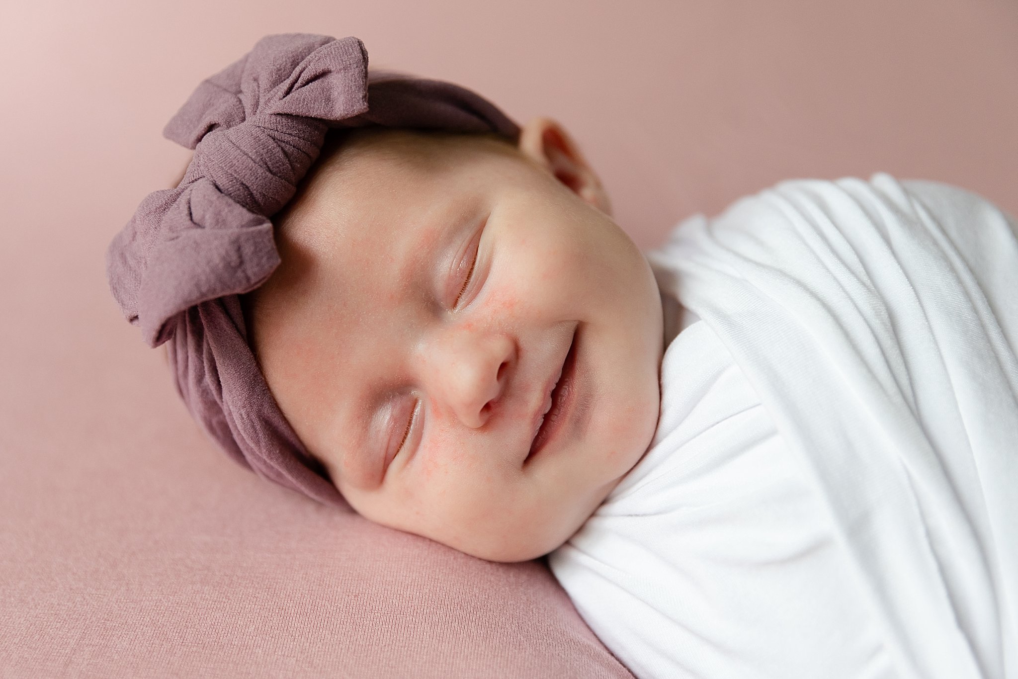 newborn girl wrapped in white on pink backdrop with purple headband