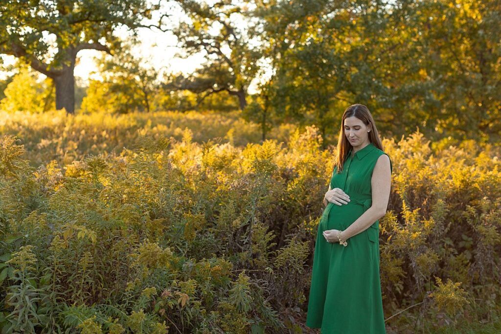 Hinsdale Maternity Photographer, Mom to be standing in a field of tall grasses and plants with the golden sun shining through the trees and tips of the brush. green maternity dress. golden hour maternity session with young kids and husband