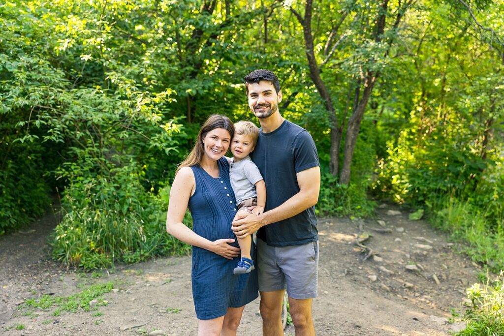 Dad and mom-to-be holding toddler boy and her belly for maternity photos on a hot summer morning